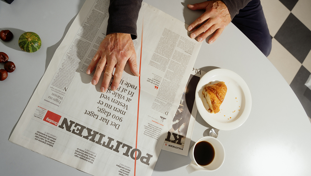 Politiken ligger på bordet ved siden af en kop kaffe og en croissant